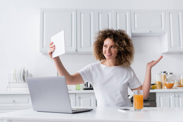 cheerful woman with wavy hair taking selfie on digital tablet near laptop in kitchen - Фото, зображення