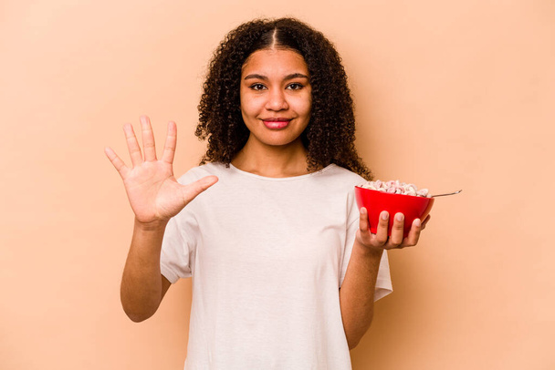Young African American woman holding a bowl of cereals isolated on beige background smiling cheerful showing number five with fingers. - Фото, изображение