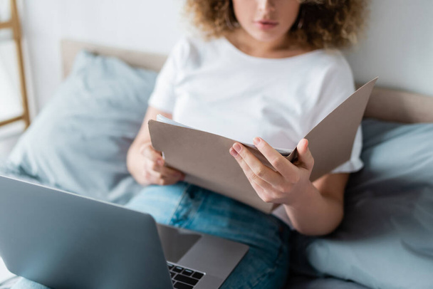 partial view of woman with folder and laptop sitting on bed on blurred background - Photo, Image
