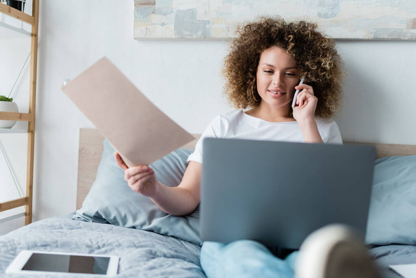 göndör nő mappával beszél okostelefon közelében laptop és digitális tabletta az ágyban - Fotó, kép