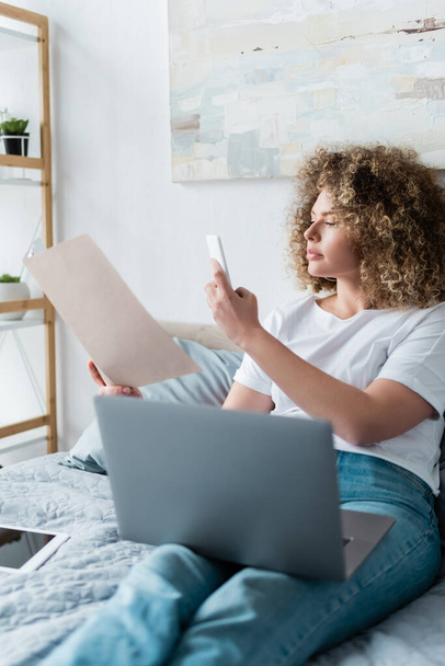 curly woman taking photo of documents on smartphone near laptop in bedroom - Foto, Imagem