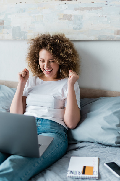 excited woman showing success gesture near laptop while sitting on bed at home - Fotografie, Obrázek