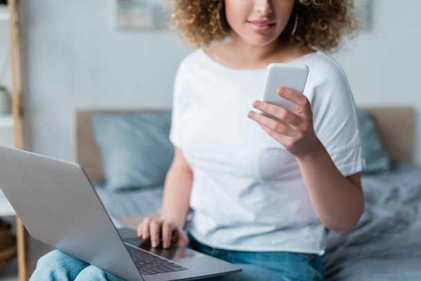 partial view of smiling woman with laptop and mobile phone in bedroom - Photo, Image