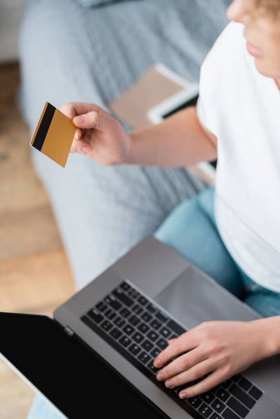 Teilansicht einer Frau mit Kreditkarte und Laptop mit leerem Bildschirm im Schlafzimmer - Foto, Bild