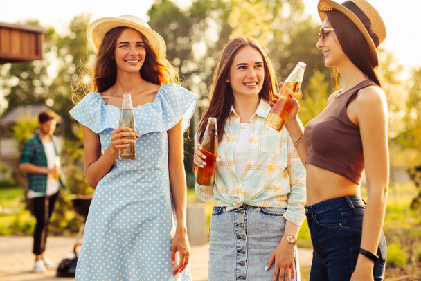 Three young beautiful smiling women in summer clothes and hats. Carefree women have a picnic on the street. Positive girls talking drinking drinks, outdoors in the park in nature - Photo, image