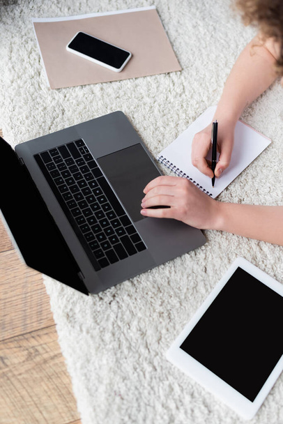 vista superior da mulher cortada escrevendo em notebook perto de dispositivos com tela em branco no chão - Foto, Imagem