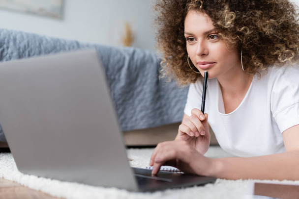 smiling woman holding pen and using laptop on floor in bedroom - Photo, Image