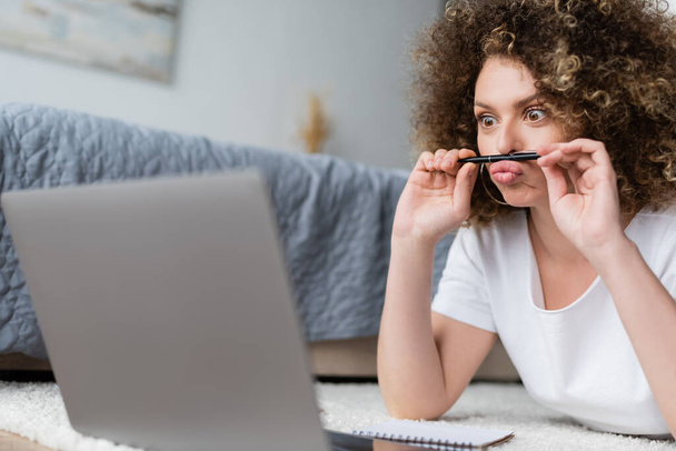 curly woman grimacing with pen while lying near laptop on floor - Foto, Imagem