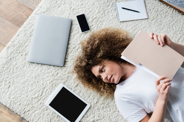 Вид сверху кудрявой женщины с папкой, лежащей на ковре рядом с устройствами и ноутбуком - Фото, изображение