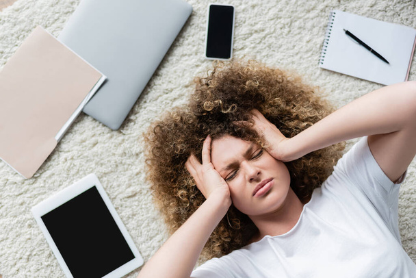 vrchní pohled vyčerpané ženy trpící bolestí hlavy v blízkosti gadgets na podlaze - Fotografie, Obrázek