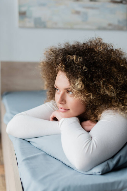 νεαρή γυναίκα με κυματιστά μαλλιά ξαπλωμένη στο κρεβάτι με σταυρωμένα χέρια και κοιτάζοντας μακριά - Φωτογραφία, εικόνα