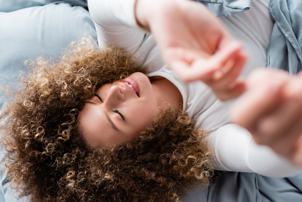 top view of smiling curly woman lying with outstretched hands on blurred foreground - Photo, Image