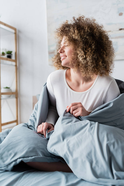 joyful woman with wavy hair sitting under grey blanket and smiling in bedroom - Zdjęcie, obraz