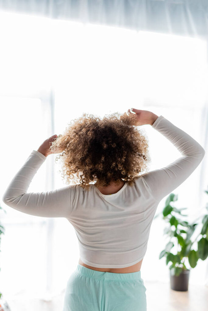 visão traseira da jovem mulher com cabelos ondulados esticando os braços de manhã em casa - Foto, Imagem