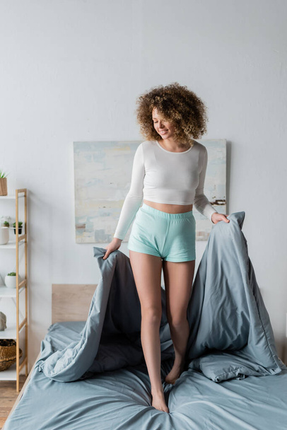 full length of curly woman in white pajamas holding grey blanket while standing on bed - Fotografie, Obrázek
