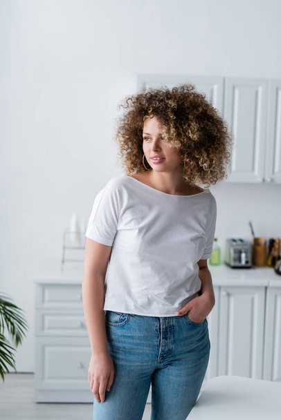 smiling woman with wavy hair standing with hand in pocket of jeans in kitchen - Fotó, kép