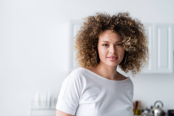 curly woman in white t-shirt smiling at camera in kitchen - Fotó, kép