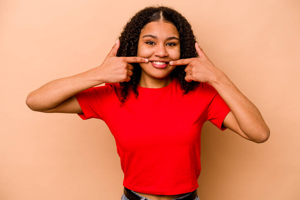 Jeune femme afro-américaine isolée sur fond beige sourit, les doigts pointant vers la bouche. - Photo, image