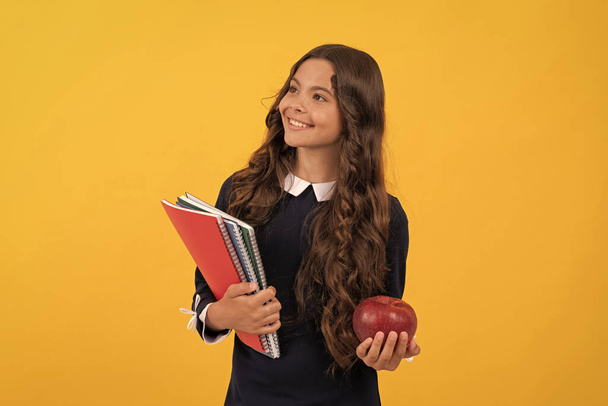 χαμογελαστός έφηβος κορίτσι κατέχουν σχολικά βιβλία και μήλο γεύμα σε κίτρινο φόντο, πίσω στο σχολείο. - Φωτογραφία, εικόνα
