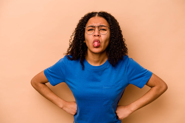 Young African American woman isolated on beige background funny and friendly sticking out tongue. - Photo, Image