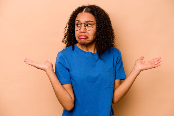 Young African American woman isolated on beige background confused and doubtful shrugging shoulders to hold a copy space. - Photo, Image