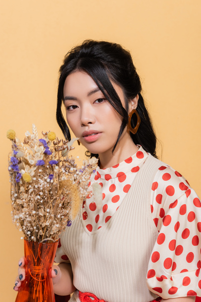 Portrait of young asian model in vintage clothes holding vase with flowers isolated on orange  - 写真・画像