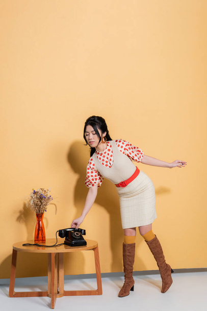 Trendy asian model holding telephone headset near flowers on coffee table on orange background - Foto, imagen
