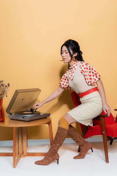 Femme asiatique élégante utilisant un tourne-disque sur fauteuil sur fond orange - Photo, image