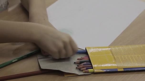 Kleine kleuter zet gekleurde potloden in doos in de buurt van blanco papier blad aan houten tafel. Kind klaar met tekenen op de kleuterschool close-up - Video
