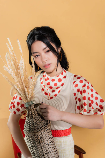 Asian woman in retro blouse and dress holding vase with spikelets on orange background - Photo, Image