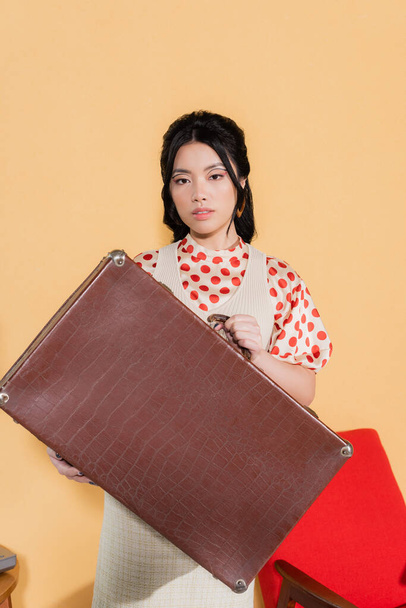 Stylish asian woman holding retro suitcase and looking at camera on orange background - Foto, afbeelding
