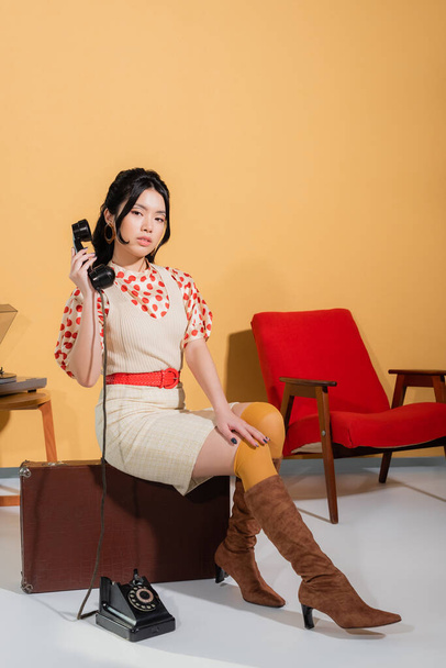 Pretty asian woman holding handset while sitting on vintage suitcase on orange background - Foto, imagen