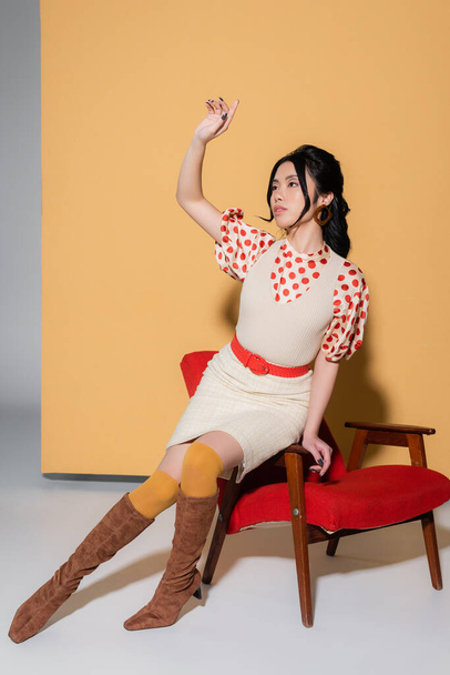 Pretty asian model in vintage clothes sitting on armchair on orange background - Photo, Image