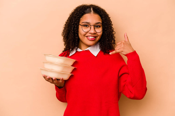 Young African American woman holding tupperware isolated on beige background showing a mobile phone call gesture with fingers. - 写真・画像