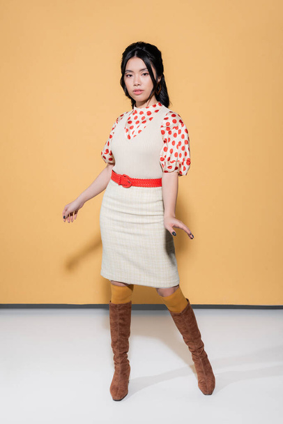 Full length of trendy asian model in retro clothes looking at camera on orange background - Photo, Image