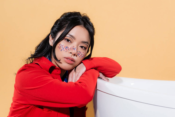 Asian model with glitter on face looking at camera near bathtub isolated on orange  - Foto, imagen