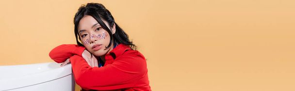 Asian model with glitter on face looking at camera near bathtub isolated on orange, banner  - Foto, afbeelding