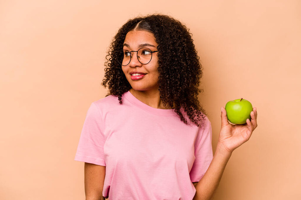 Joven mujer afroamericana sosteniendo una manzana aislada sobre fondo beige mira a un lado sonriente, alegre y agradable. - Foto, imagen