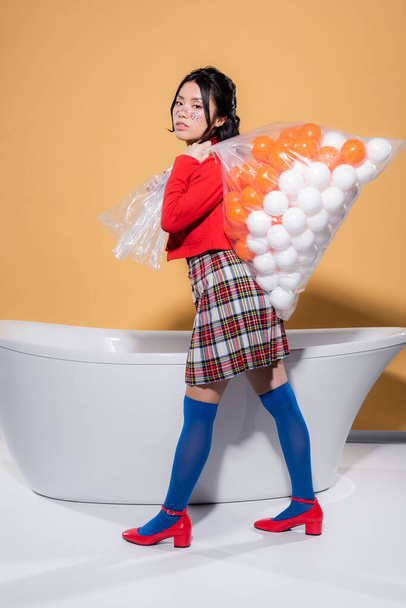 Trendy asian woman in vintage clothes holding plastic bag with balls near bathtub on orange background - 写真・画像