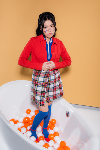 Elegante mulher asiática olhando para a câmera enquanto em pé na banheira com bolas no fundo laranja - Foto, Imagem