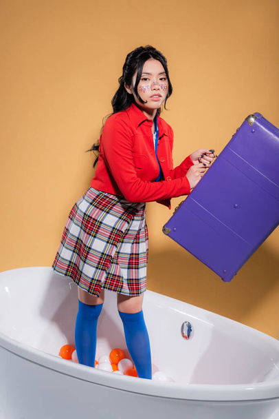 Fashionable asian woman with glitter on face holding retro suitcase in bathtub with balls on orange background - Foto, afbeelding