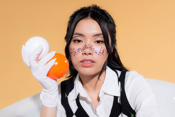 Portrait of young asian model with glitter on face holding balls and looking at camera in bathtub isolated on orange  - Photo, Image