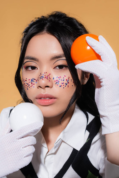 Portrait de modèle asiatique avec visage pailleté tenant des boules et regardant la caméra isolée sur orange  - Photo, image