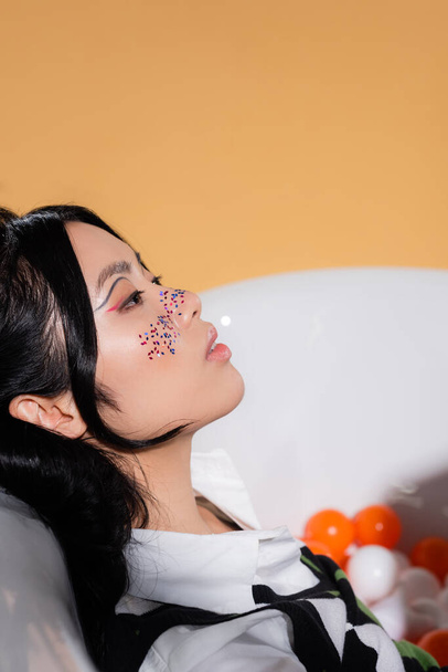 Vista lateral da mulher asiática na moda com maquiagem brilho sentado na banheira turva com bolas isoladas na laranja  - Foto, Imagem