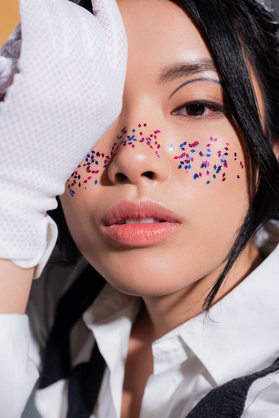 Portrait of stylish asian model with glitter on face looking at camera isolated on orange  - Photo, image