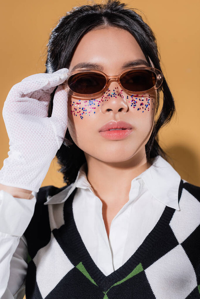 Portrait of stylish asian model holding sunglasses on orange background - Foto, afbeelding