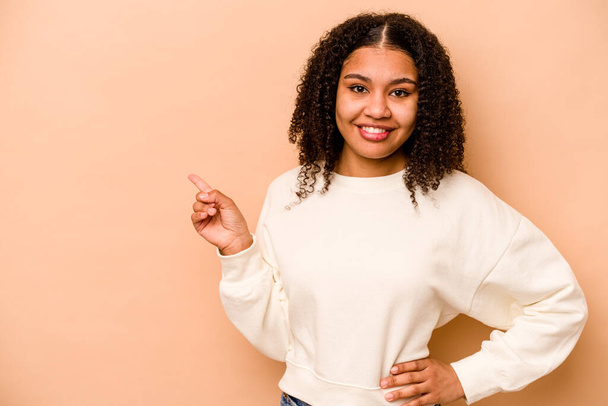 Joven mujer afroamericana aislada sobre fondo beige sonriendo alegremente señalando con el dedo índice de distancia. - Foto, imagen