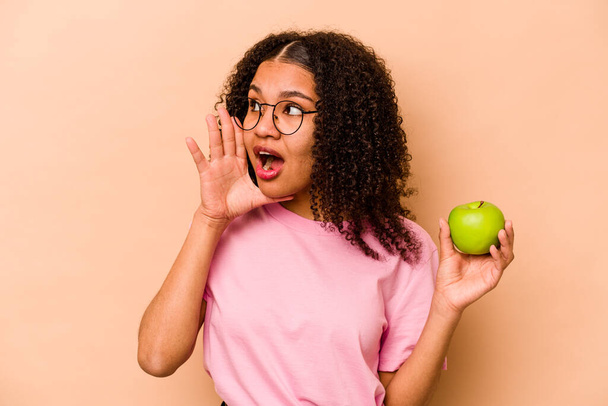 Молодая афроамериканка держит яблоко на бежевом фоне крича и держа ладонь у открытого рта. - Фото, изображение