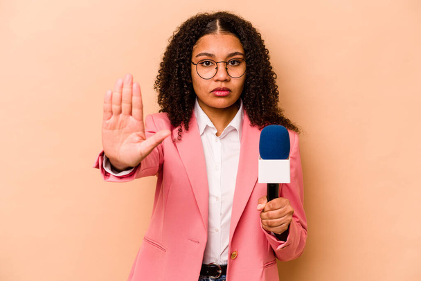 Joven presentadora de televisión afroamericana aislada sobre fondo beige de pie con la mano extendida mostrando señal de stop, lo que le impide. - Foto, Imagen