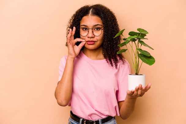 Young African American woman holding a plant isolated on beige background with fingers on lips keeping a secret. - Photo, Image